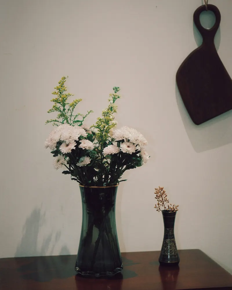 Dunkler Holztisch mit einer Vase voller heller Wiesenblumen vor einer weißen Wand